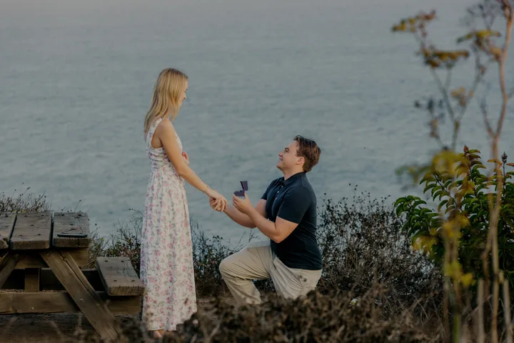 Jack kneeling with a ring box as Kayley stands beside him above the ocean