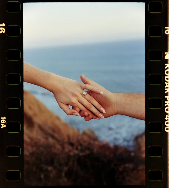 Film-framed detail of Kayley and Jack holding hands above the ocean